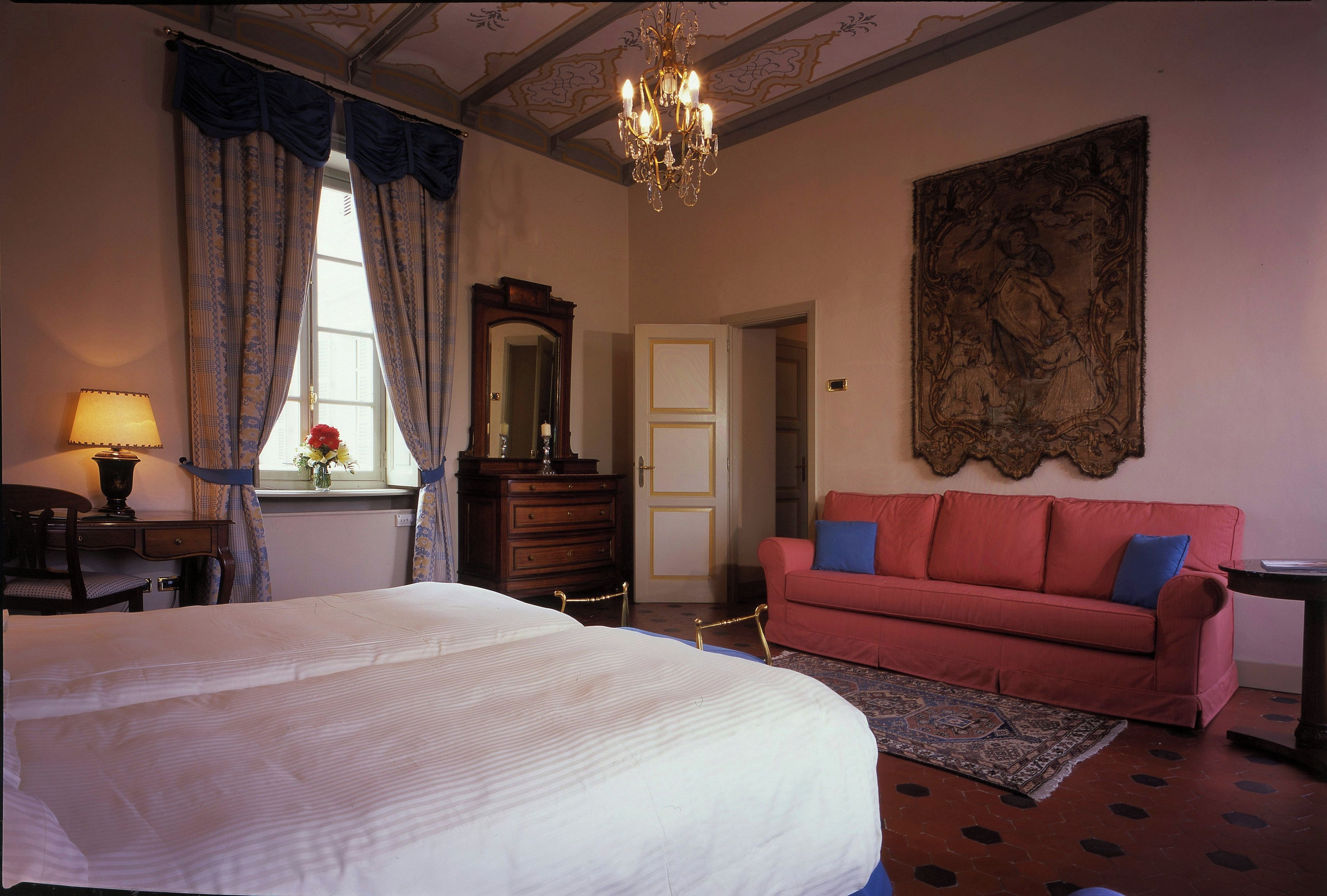 A bedroom with a large bed, a red sofa, a chandelier, a large painting, and a wooden dresser.