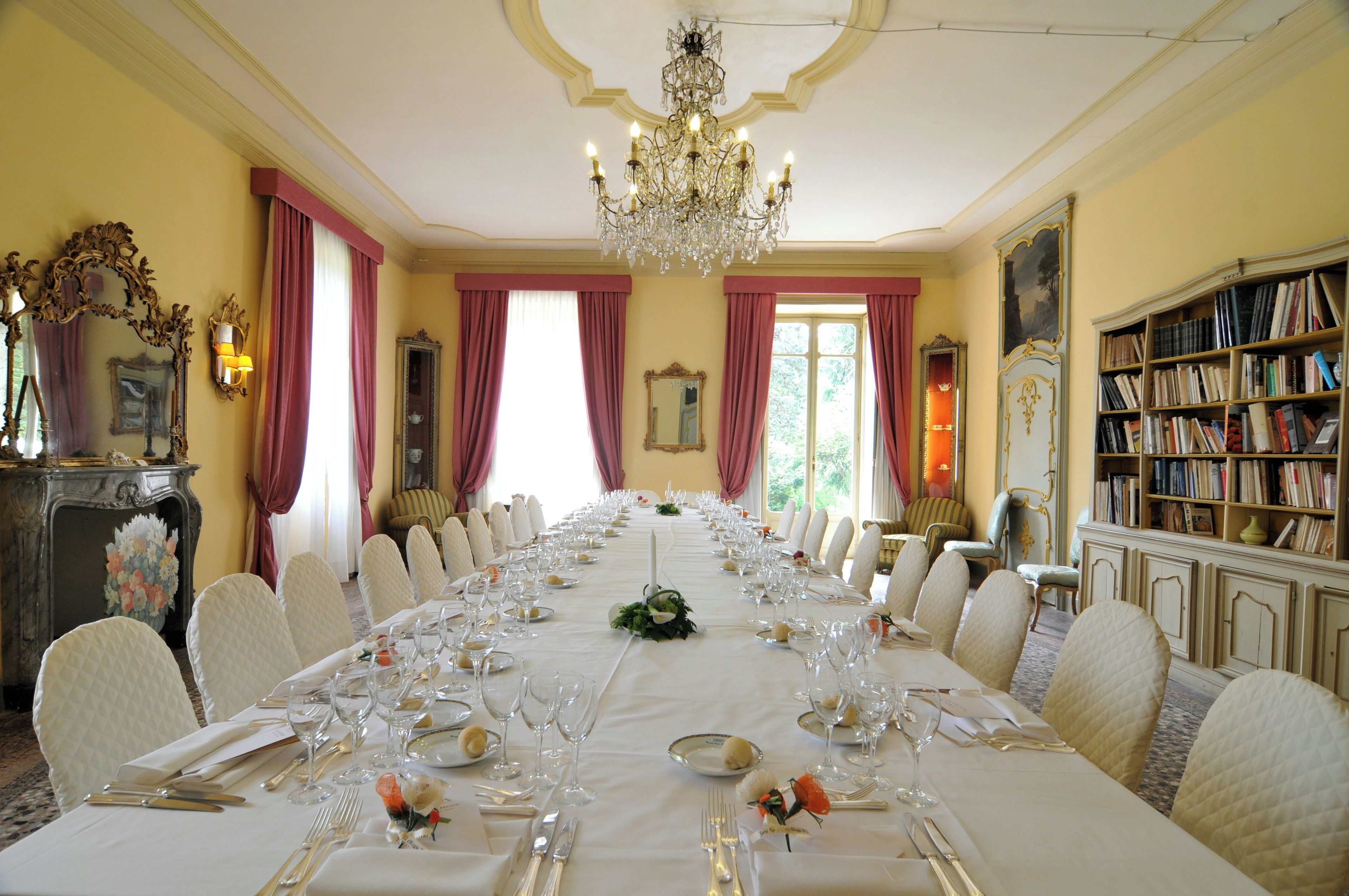 Ein stilvoller Speisesaal mit einem langen Tisch, gedeckt für eine Mahlzeit, Kronleuchtern und einer Bücherwand.