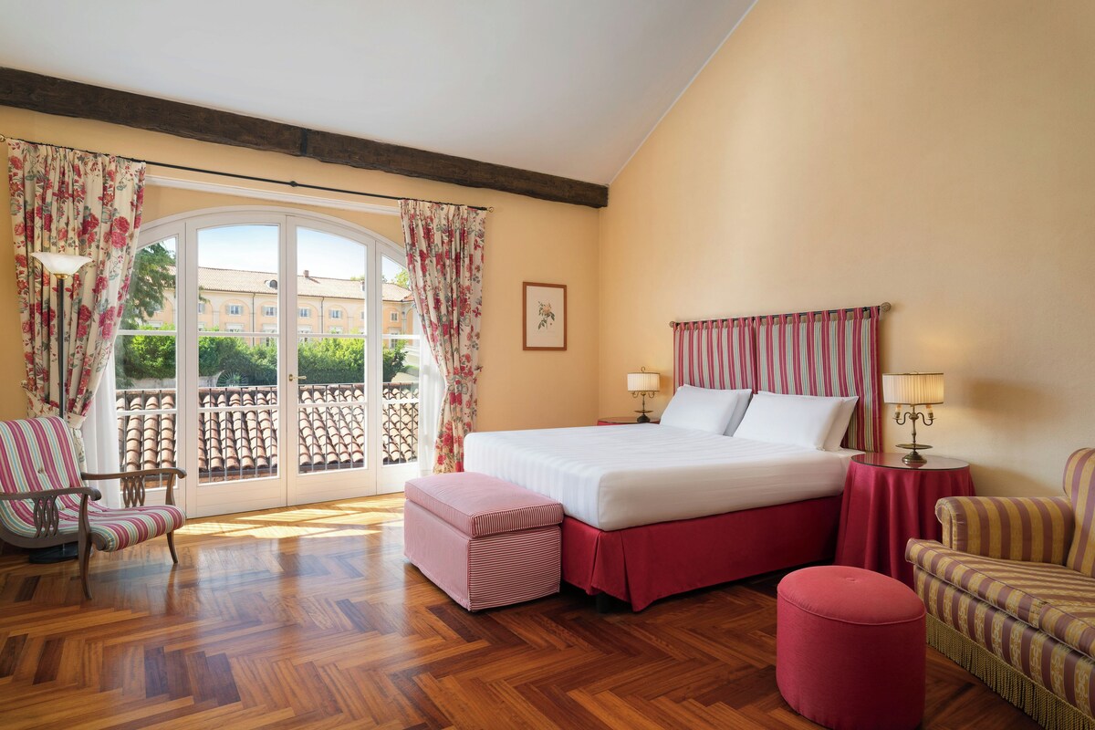 Una habitación de hotel con cama, dos sillas, una mesita y un balcón con cortinas.