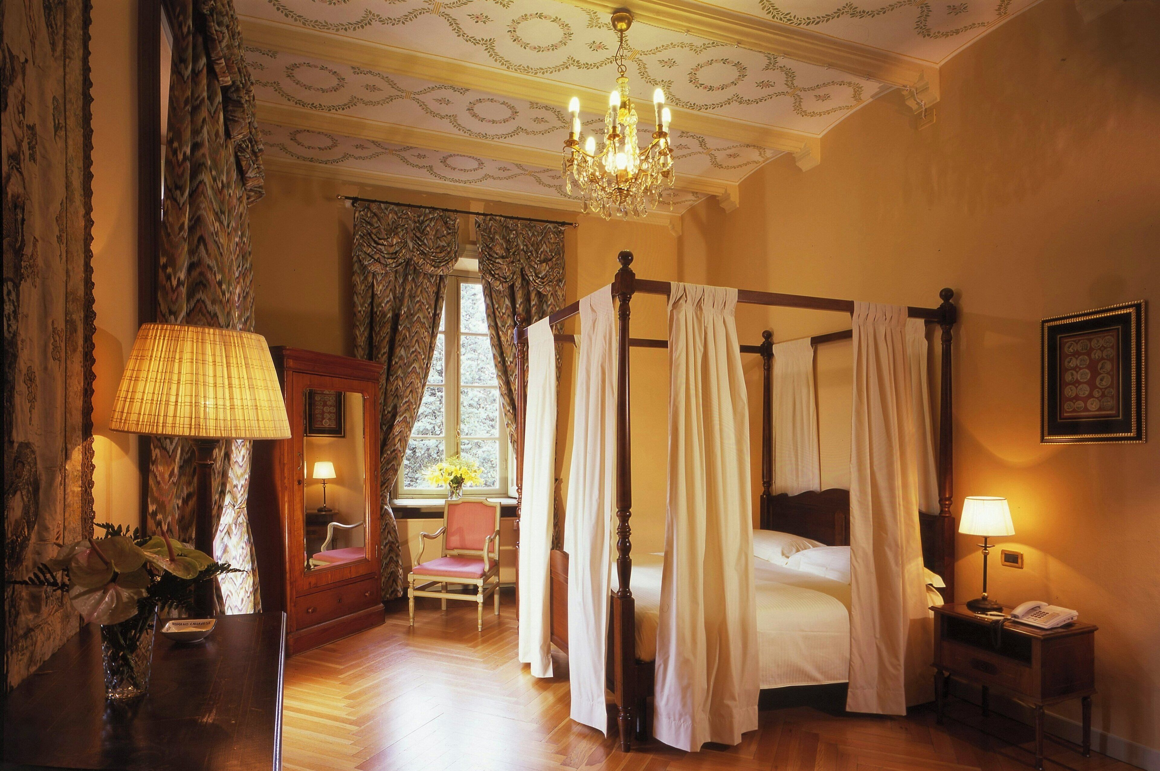 A four-poster bed with a canopy, a sitting chair, a bedside table with a lamp, and a window with curtains.