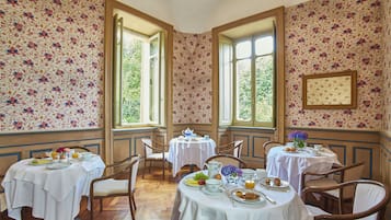 Ein Essbereich mit floraler Tapete, mehreren Tischen, Stühlen und großen Fenstern.