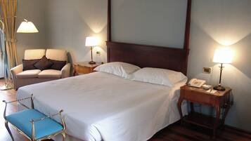 A neatly made bed with white linens, a wooden headboard, two bedside lamps, a chair with a cushion, and a small bedside table with a phone.