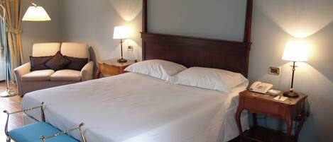 A neatly made bed with white linens, a wooden headboard, two bedside lamps, a chair with a cushion, and a small bedside table with a phone.