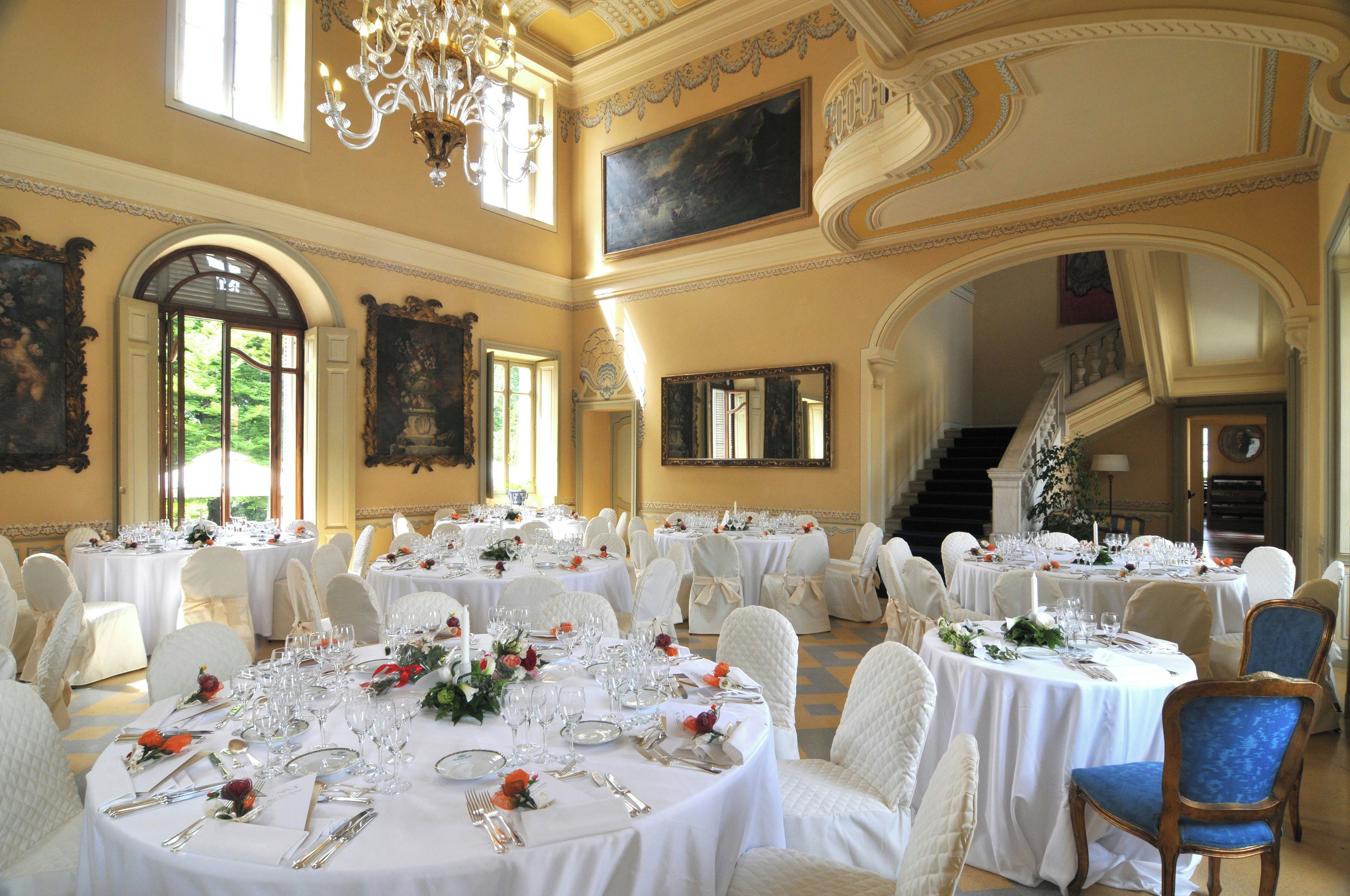 A grand dining hall with multiple round tables set for a formal event, featuring chandeliers and classical architecture.