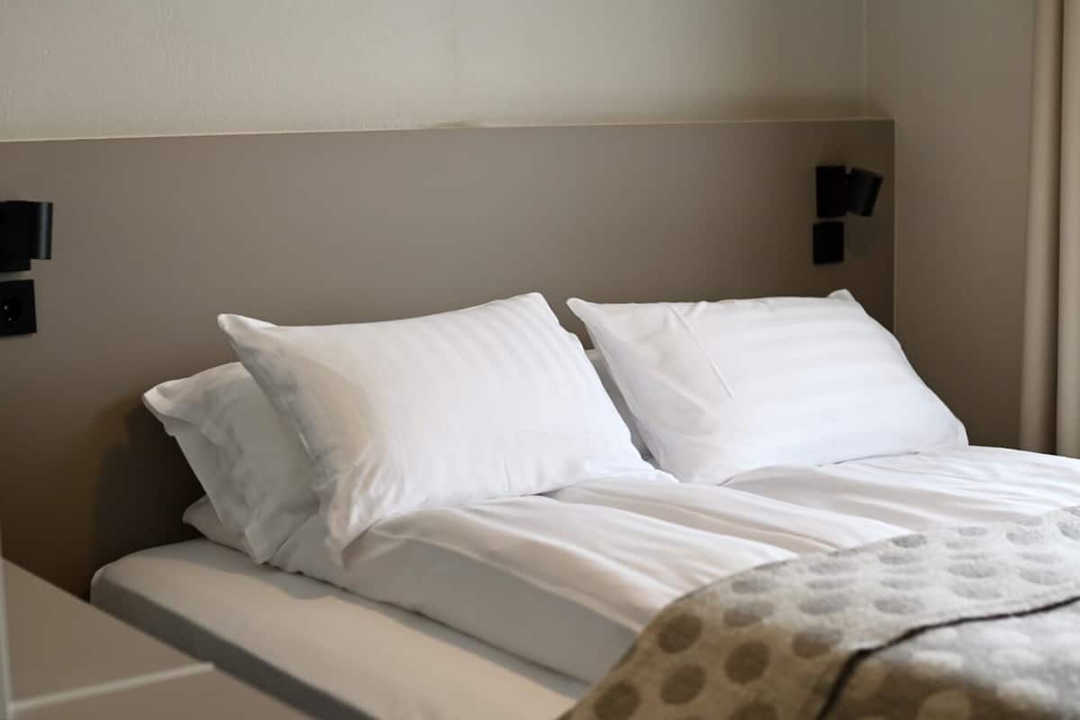 A neatly made bed with white linens and two pillows, a headboard with wall-mounted lights, and a partial view of a patterned blanket.