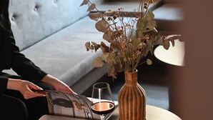 Una persona leyendo un libro en una mesa redonda, con una copa de vino, un jarrón con flores secas y un sofá capitoné al fondo.