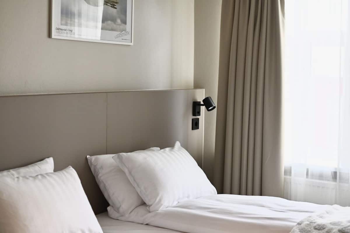 A hotel room with a bed, a headboard, a wall-mounted lamp, and a window with curtains.