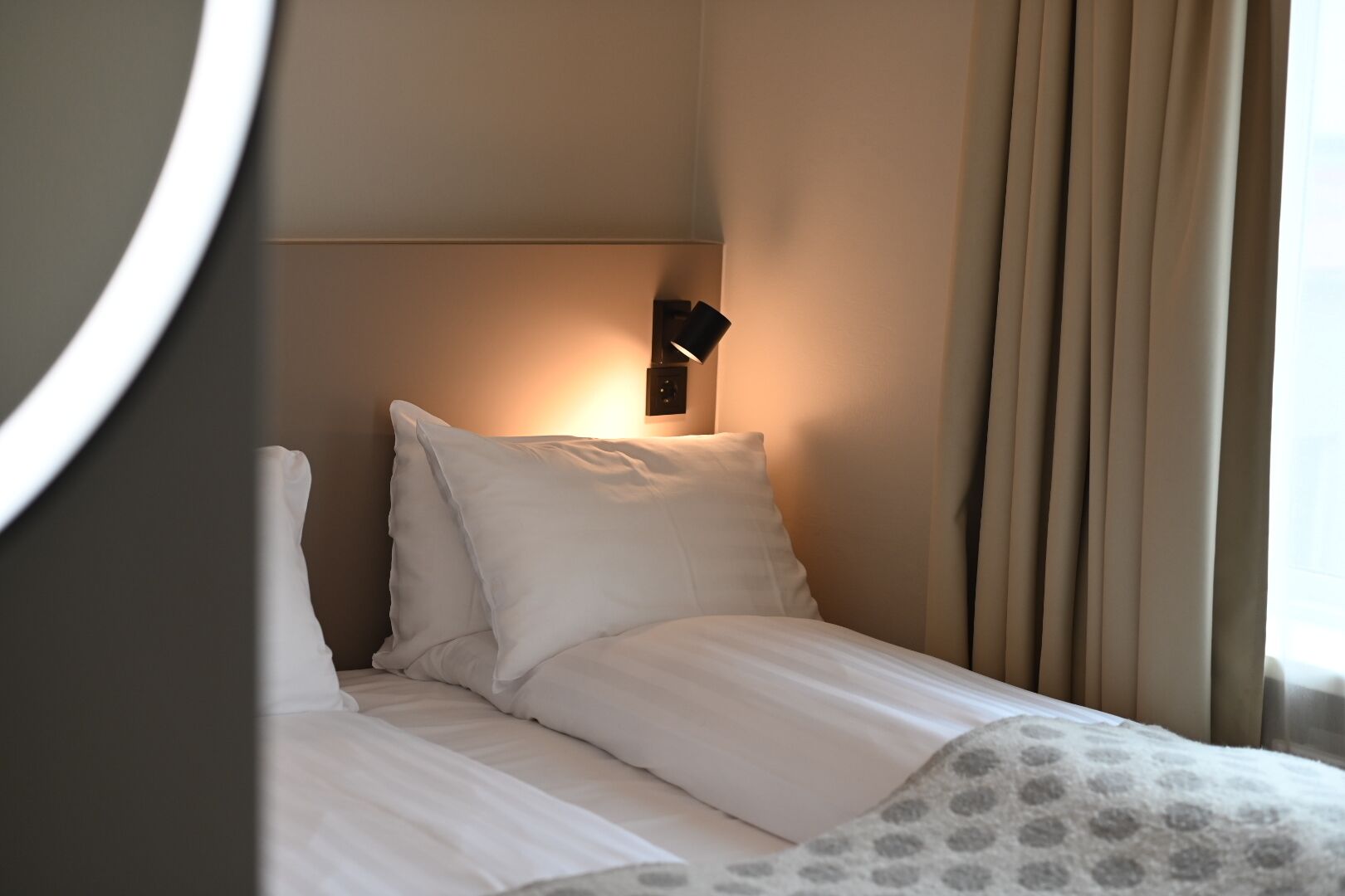 Ein Hotelzimmer mit einem ordentlich bezogenem Bett, einer Wandleuchte und einem Fenster mit Vorhängen.