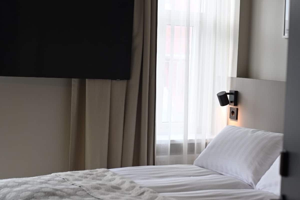 A bedroom with a bed, a TV mounted on the wall, a window with curtains, and a bedside lamp.