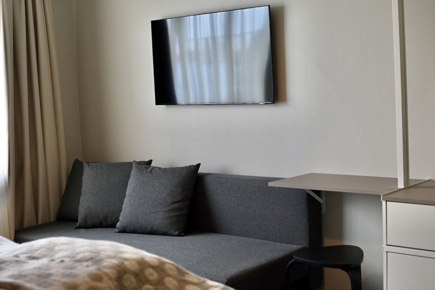 A modern hotel room with a grey sofa, a flat-screen TV mounted on the wall, a small desk, and a bedside table.