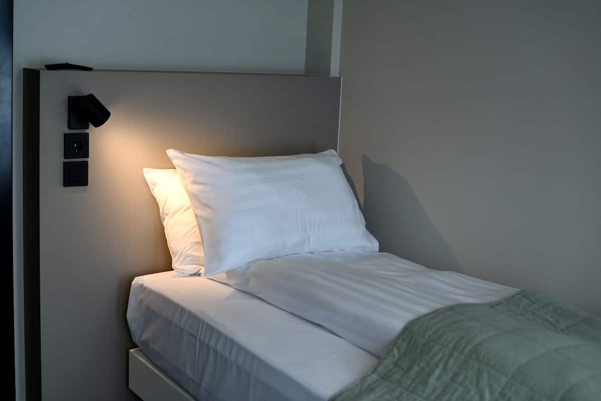 A single bed with white bedding and a green blanket, a wall-mounted light, and a small black object on the wall.