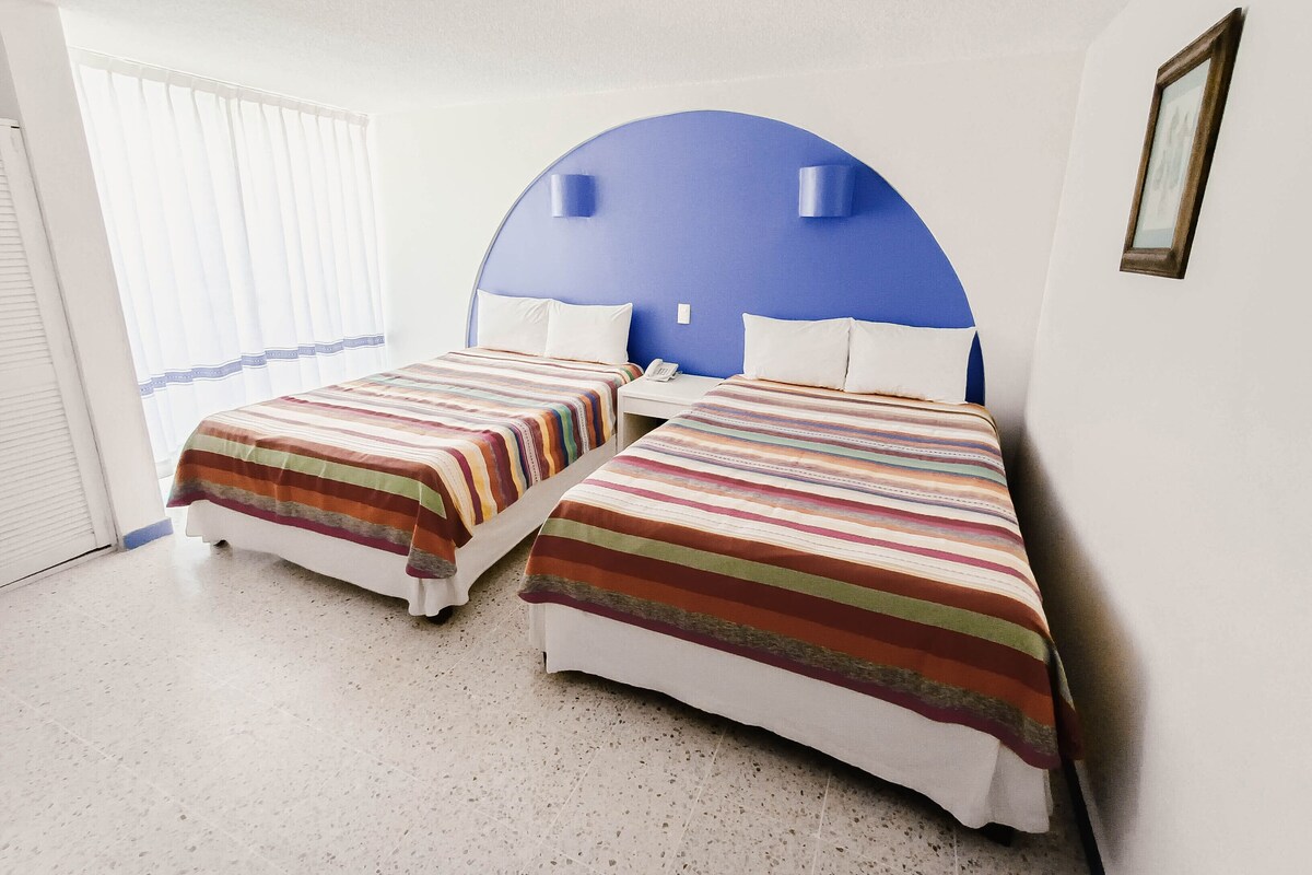 Une chambre d’hôtel avec deux lits, une grande fenêtre avec des stores et un mur d’accent bleu.