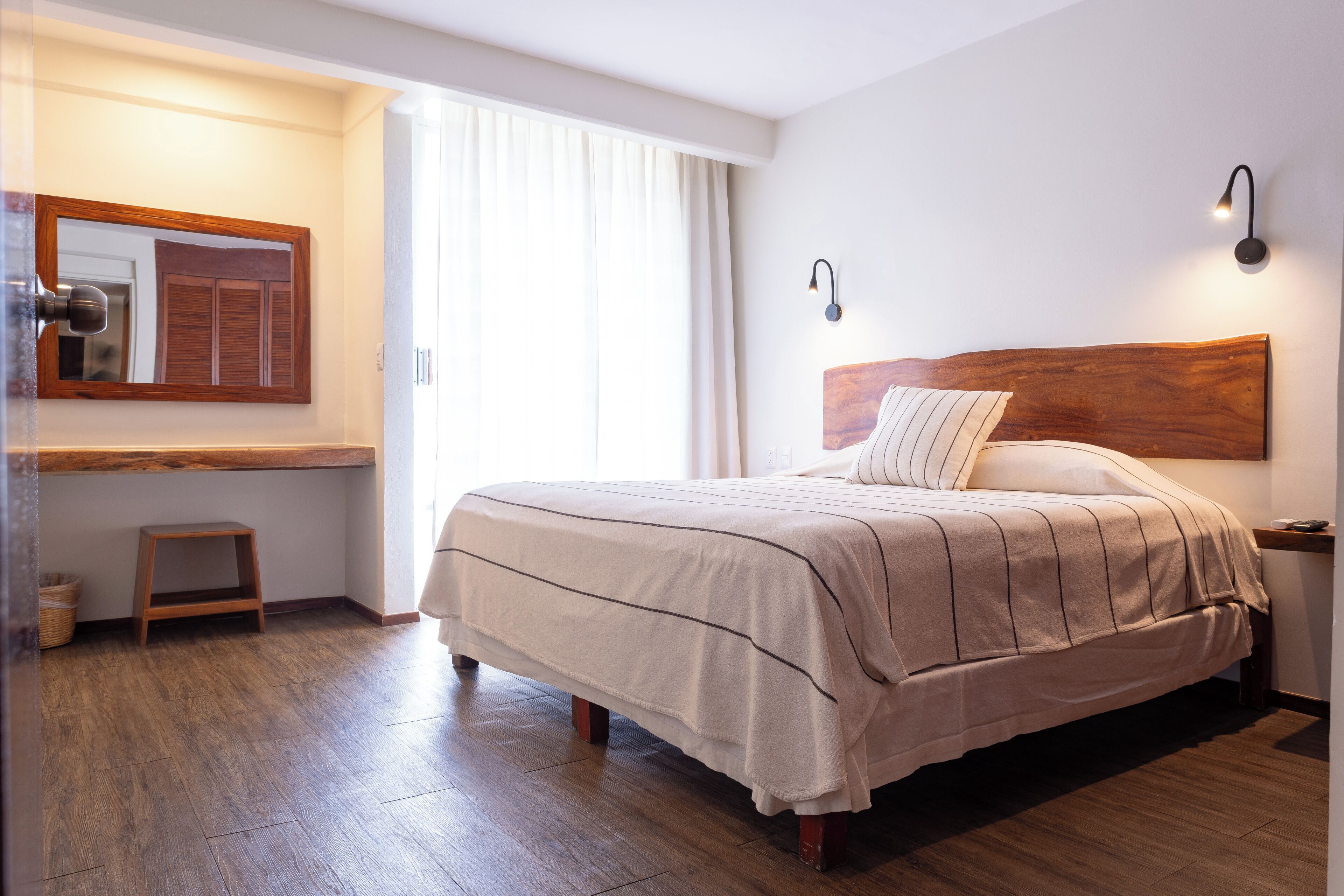 Un dormitorio ordenado con una cama grande, un taburete de madera, una mesita de noche y un espejo adosado a la pared.