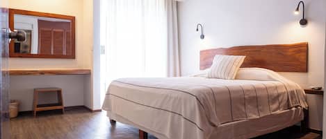 Un dormitorio ordenado con una cama grande, un taburete de madera, una mesita de noche y un espejo adosado a la pared.