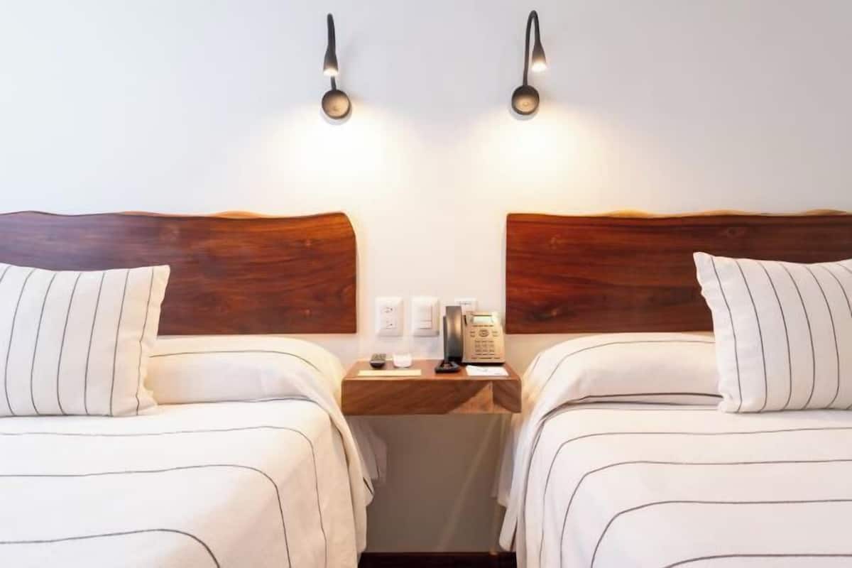 Hotellihuone, jossa on kaksi sänkyä, puiset päätyosat, yöpöydät lampuilla sekä pieni työpöytä, jossa on puhelin ja kuppi.