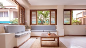 Uma sala de estar moderna com sofá, mesa de centro de madeira e vista para uma piscina e palmeiras através de amplas janelas com venezianas de madeira.