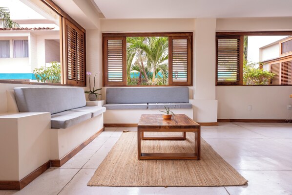 Un salon moderne avec un canapé, une table basse en bois et une vue sur une piscine et des palmiers, visible à travers de grandes fenêtres dotées de volets en bois.