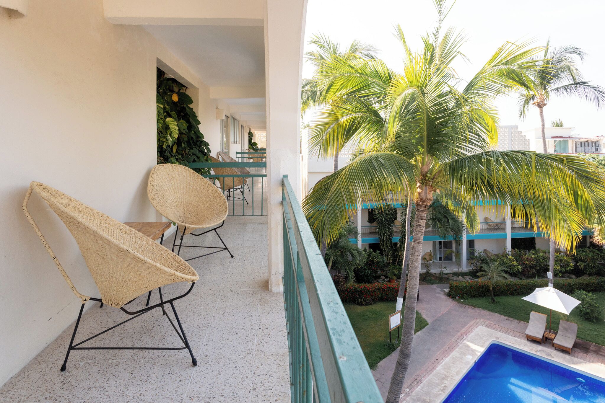 A balcony with wicker chairs, a pool, and a view of a building with a balcony and palm trees.