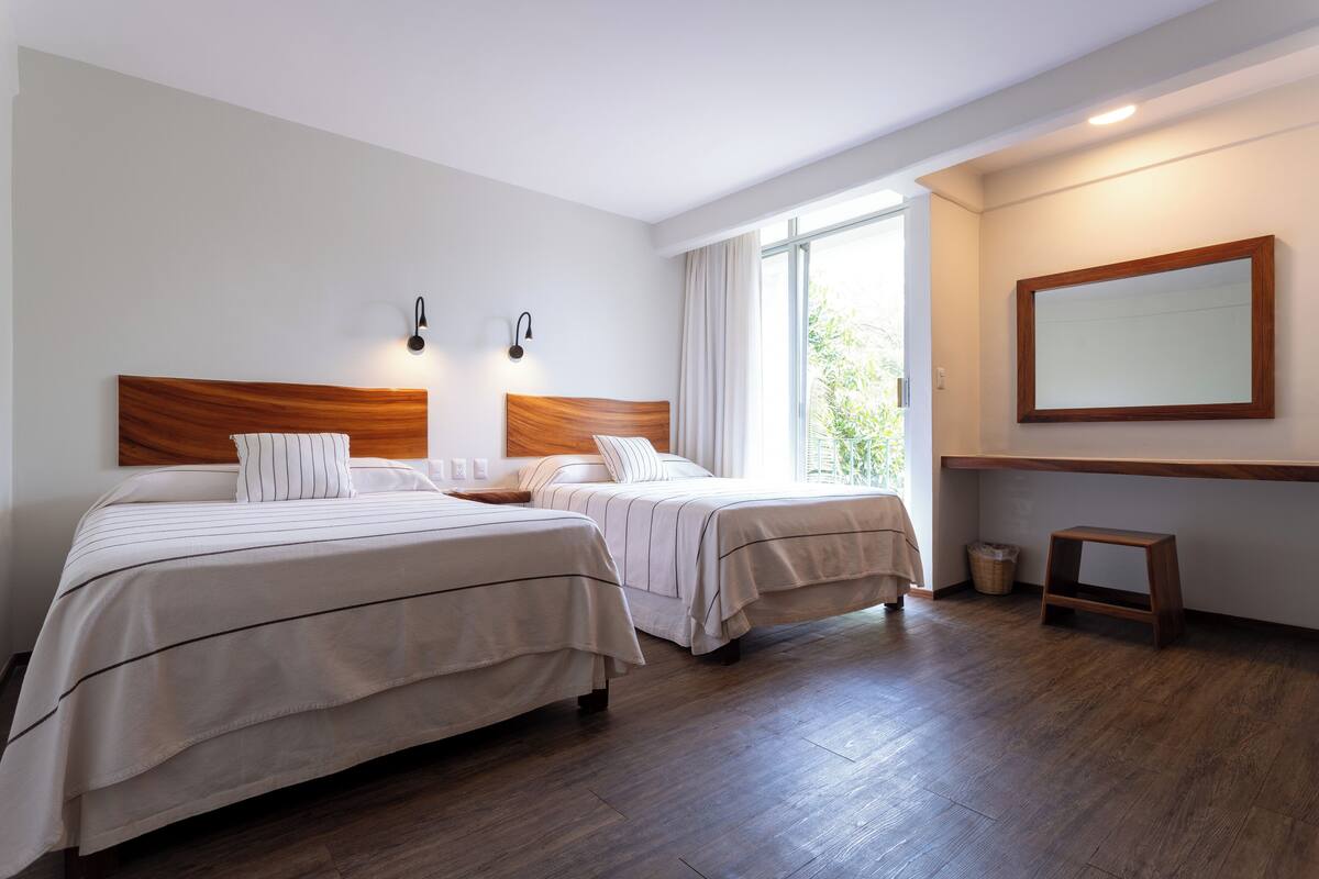 Hotellihuone, jossa on kaksi sänkyä, puinen pääty, suuri ikkuna verhoilla, puulattia ja seinälle kiinnitetty peili.