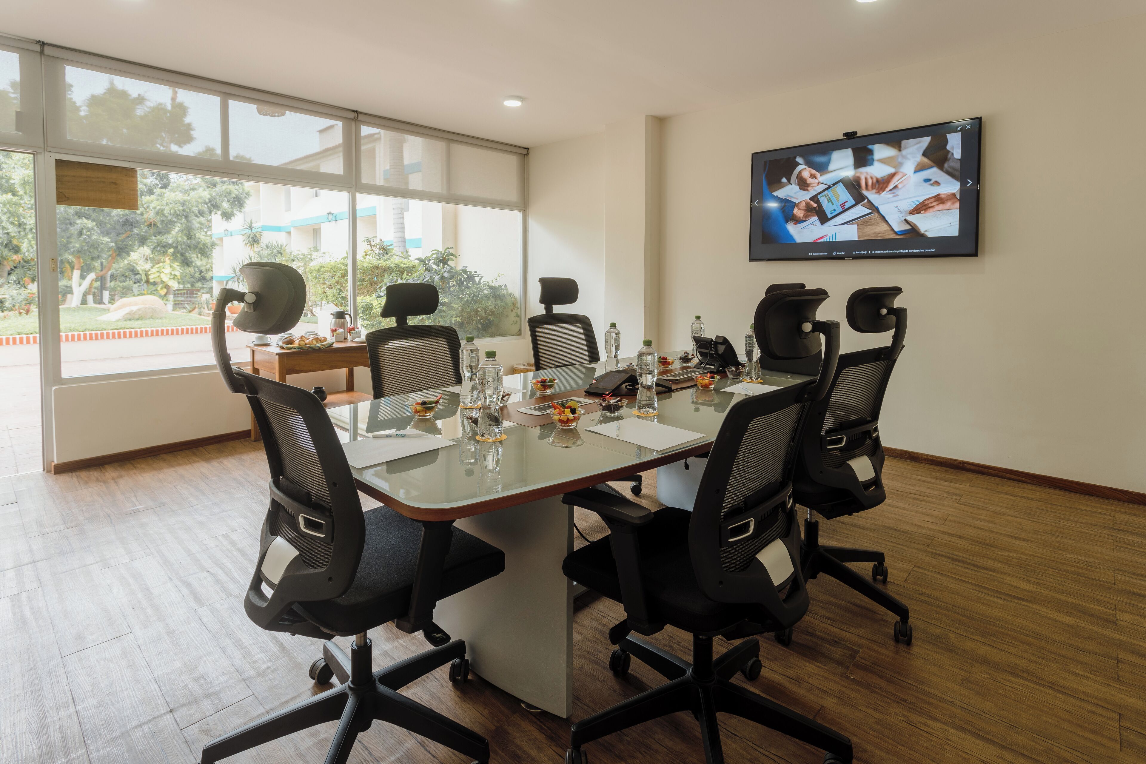 Un salón de conferencias con una mesa grande, cuatro sillas, un televisor en la pared y vistas al exterior a través de amplias ventanas.