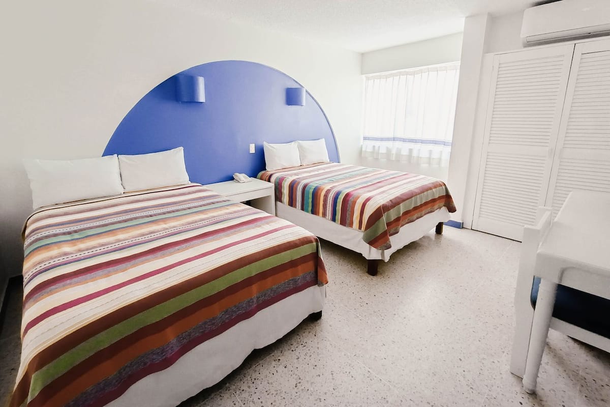 A hotel room with two beds, a blue accent wall, a white wardrobe, and a window with white curtains.