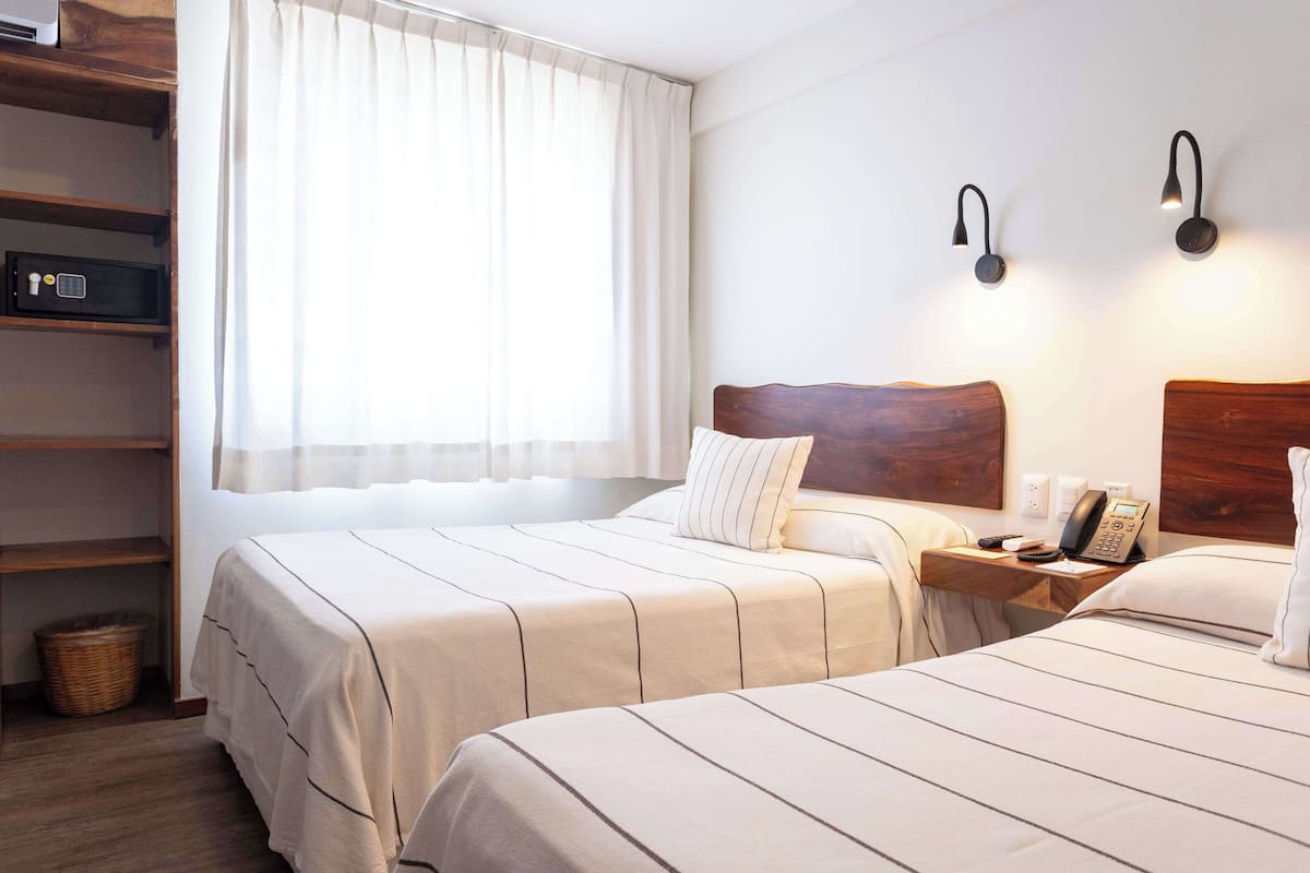 Hotellihuone, jossa on kaksi sänkyä, puinen pääty, yöpöytä, jossa on puhelin, ja ikkuna, jossa on verhot.