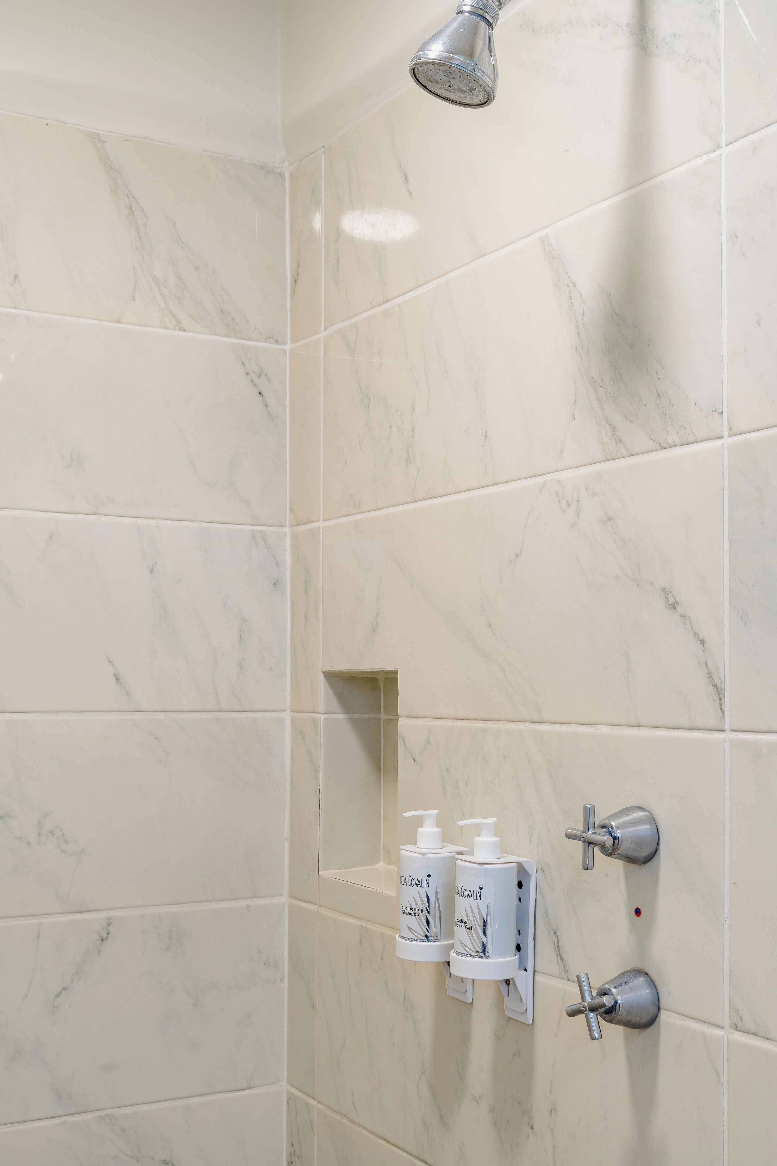 Une salle de bain carrelée de marbre, avec une pomme de douche et deux distributeurs de savon.