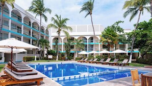 Zona de piscina con sillones de playa y sombrillas, rodeada por un edificio de varios pisos con balcones.