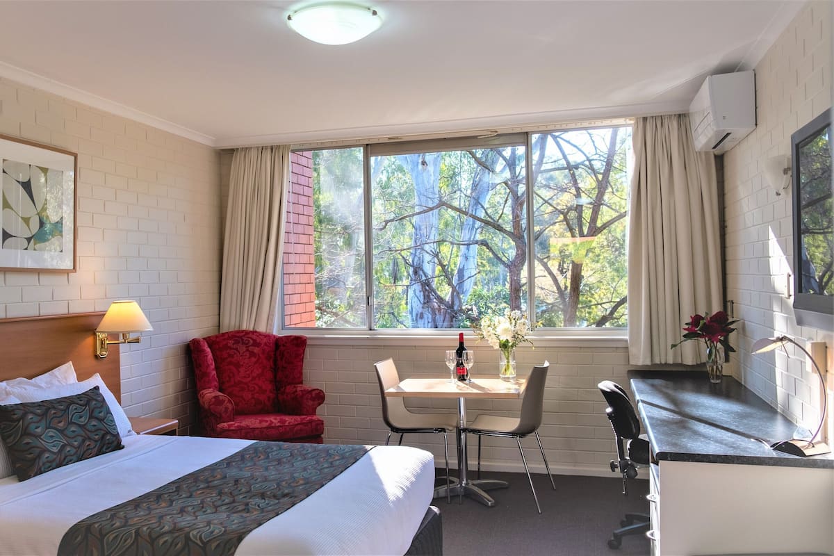 A hotel room with a bed, a desk, a chair, a table, and a window with a view of trees.