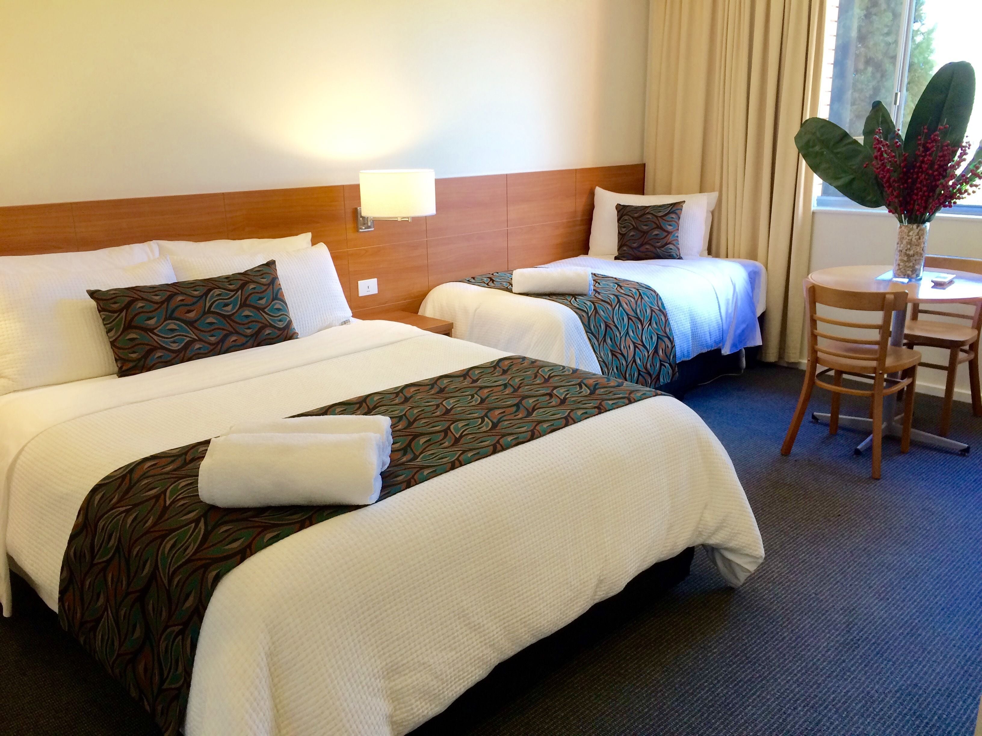 Una habitación de hotel con dos camas, una mesita pequeña, sillas, una lámpara y una ventana con cortinas.