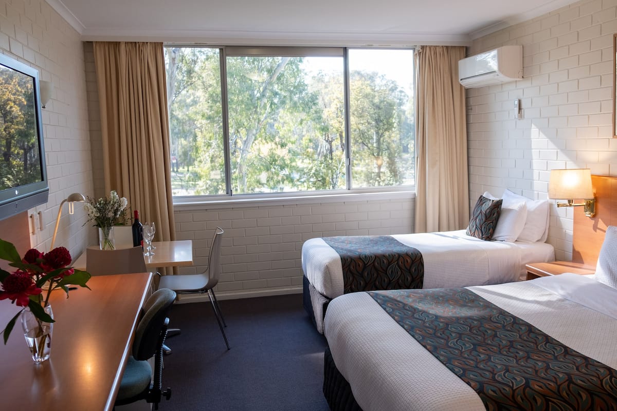 A hotel room with two beds, a desk with a chair, a window with curtains, an air conditioning unit, and a framed picture on the wall.