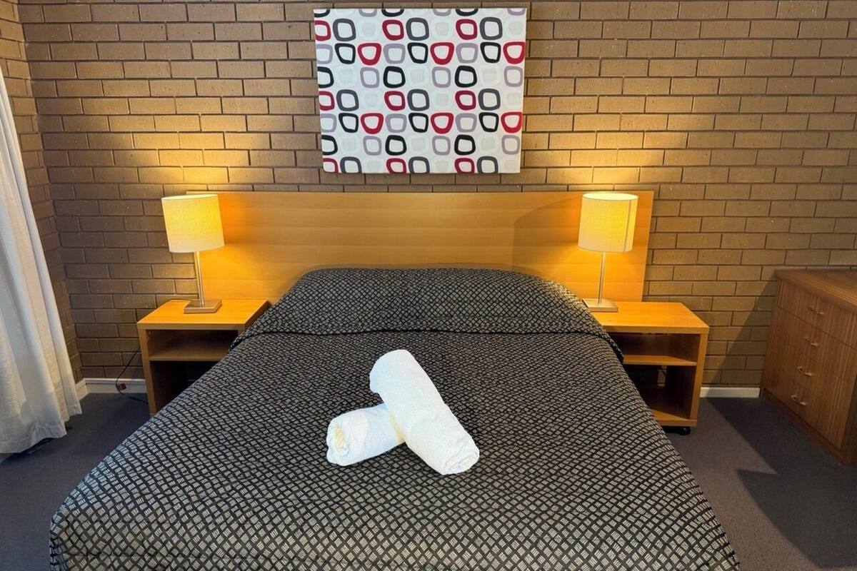 A hotel room with a bed, two bedside lamps, a headboard, and a wall-mounted artwork.