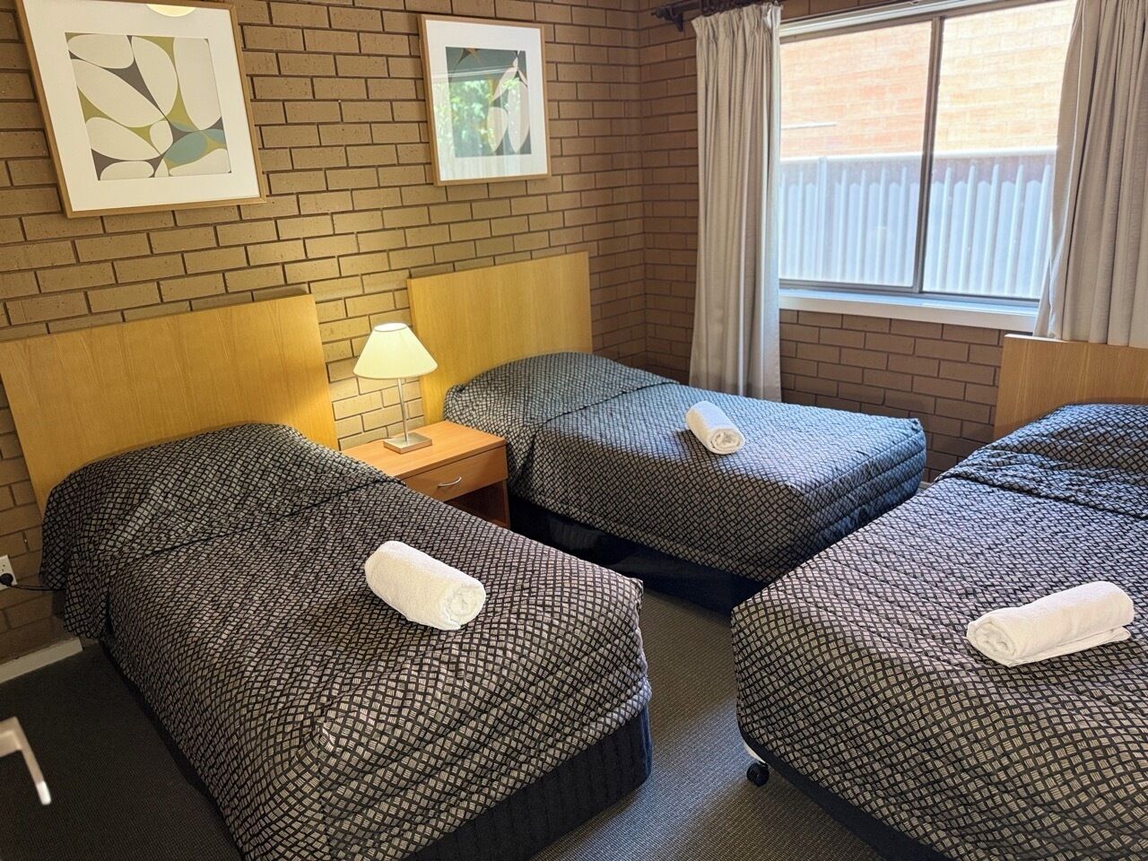 Habitación de hotel con tres camas, cada una con una colcha estampada y una toalla enrollada. Hay una mesita de noche con una lámpara entre las camas, y la habitación tiene paredes de ladrillo y una ventana con cortinas.