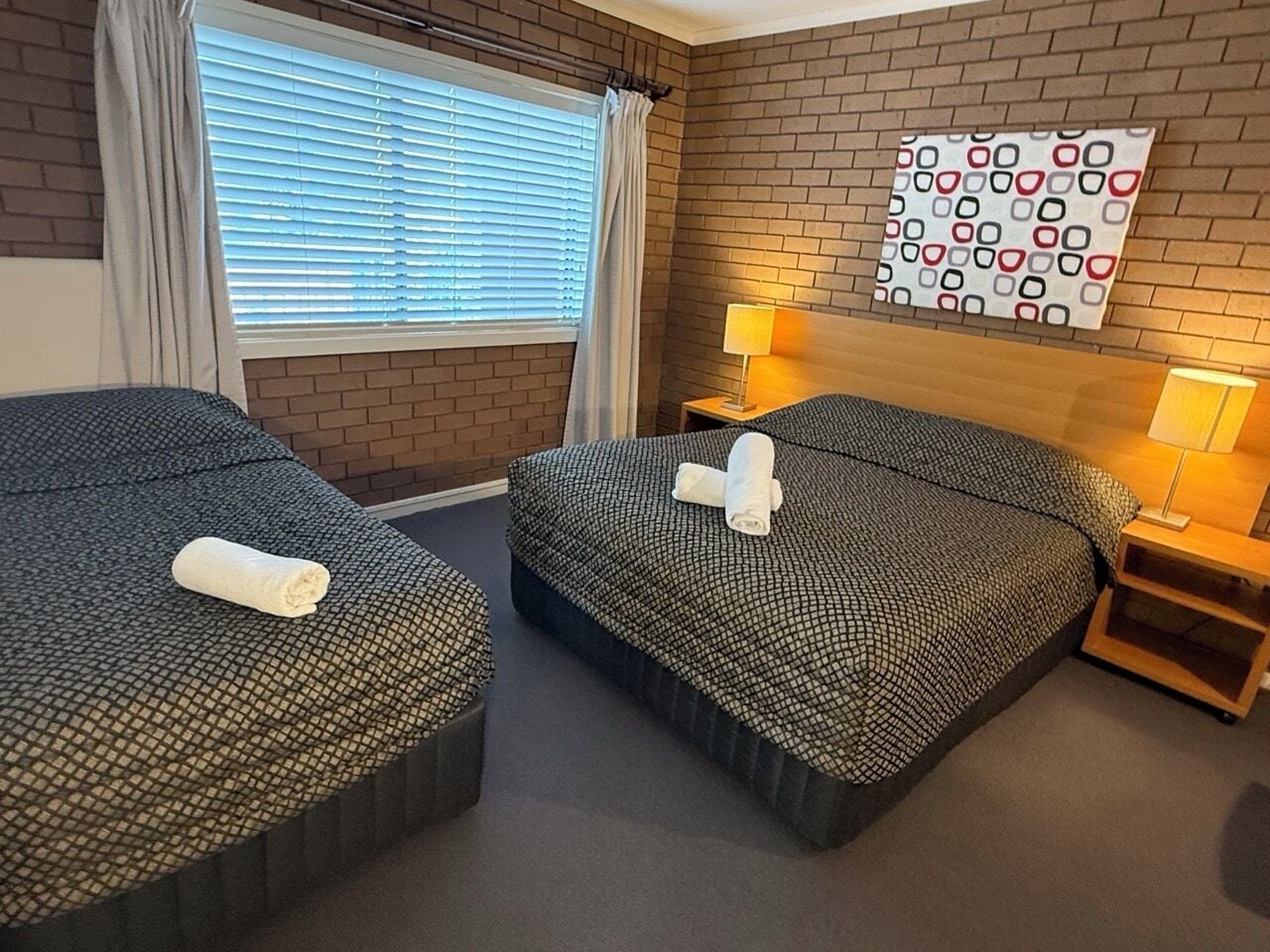 Habitación de hotel con dos camas, una ventana con persianas, un muro de ladrillo, un adorno mural estampado y lámparas de noche.