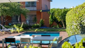 Zona de piscina con sillones de descanso y una mesa, rodeada por un edificio de ladrillo y vegetación.