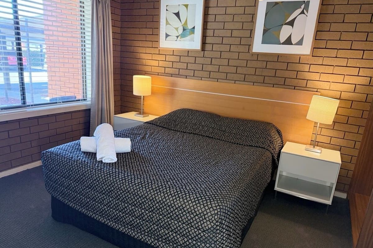 A hotel room with a large bed, two bedside tables, a window with blinds, and two framed artworks on the wall.