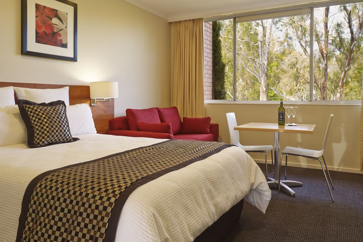 A hotel room with a large bed, a red sofa, a small table with a bottle and glasses, and a window with a view of trees.