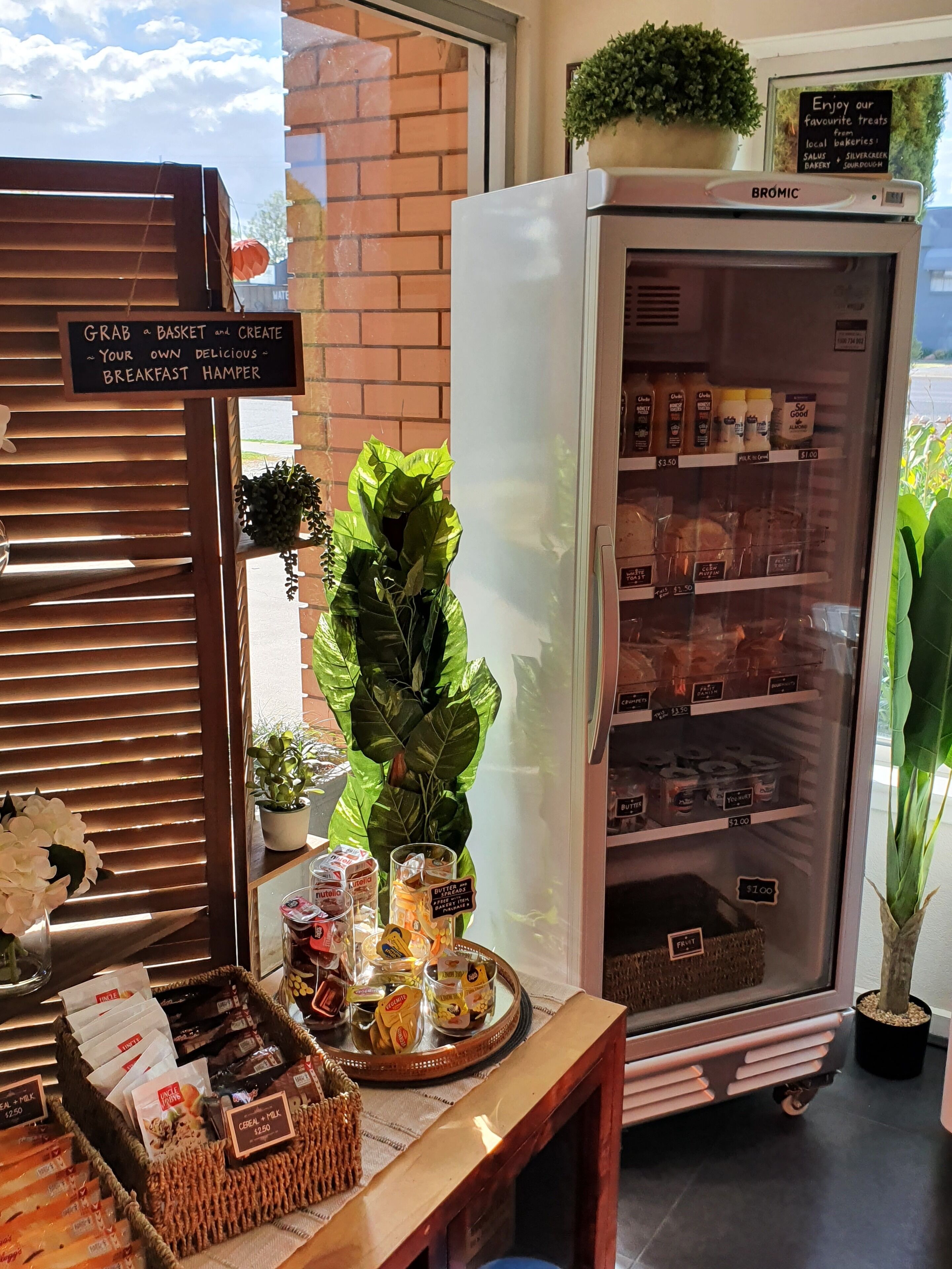 A breakfast hamper stand with a sign, a refrigerator stocked with bakery items, and a display of snacks and drinks.