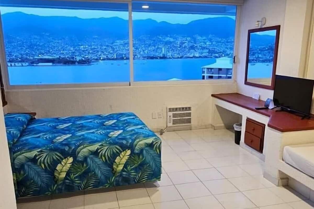 Una habitación con cama, escritorio, silla, televisor y un amplio ventanal con vista a la ciudad y montañas.