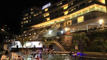 Un hotel con área de piscina por la noche, que presenta varios niveles y mobiliario exterior.