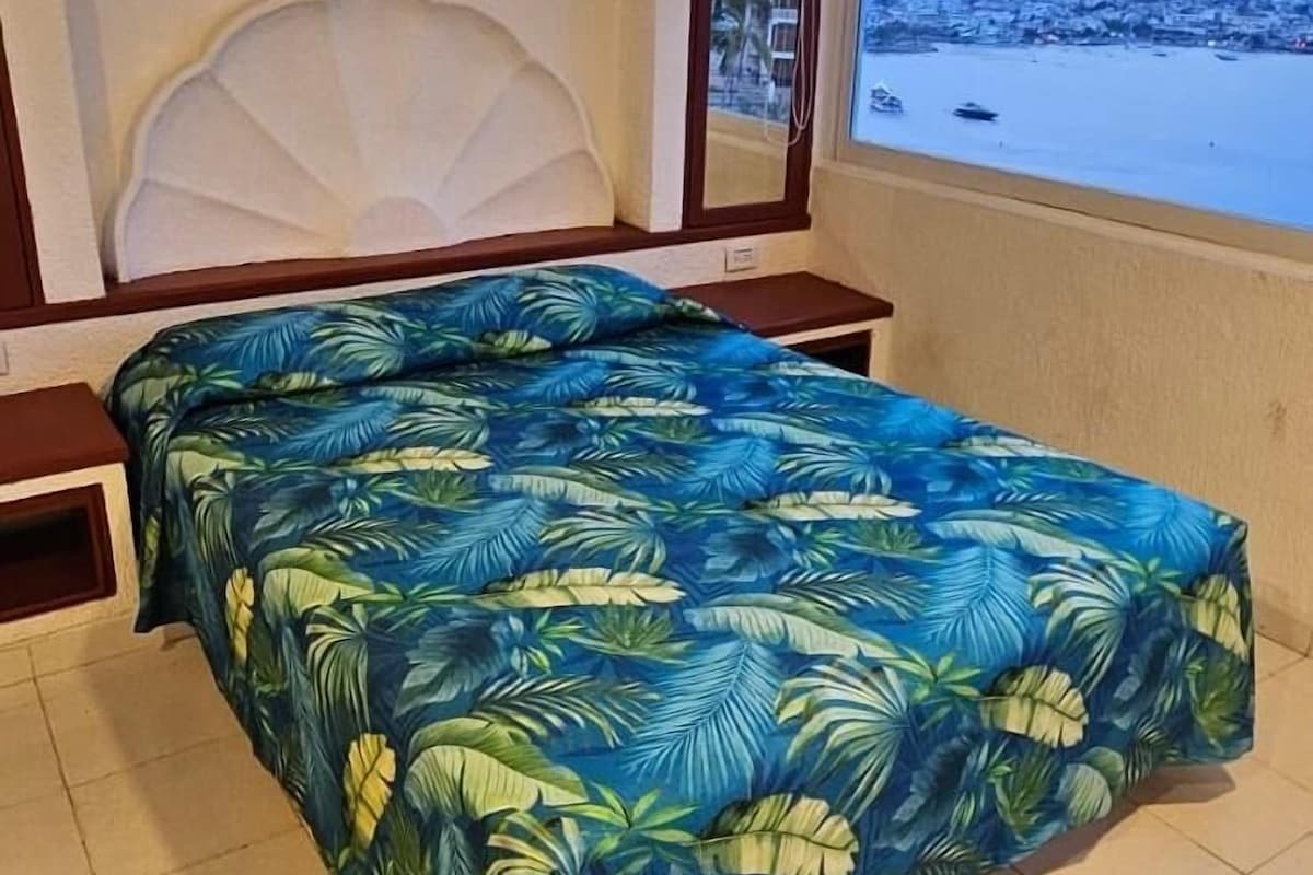 A bed with a tropical leaf pattern bedspread, a headboard, and a window with a view of a coastal area.
