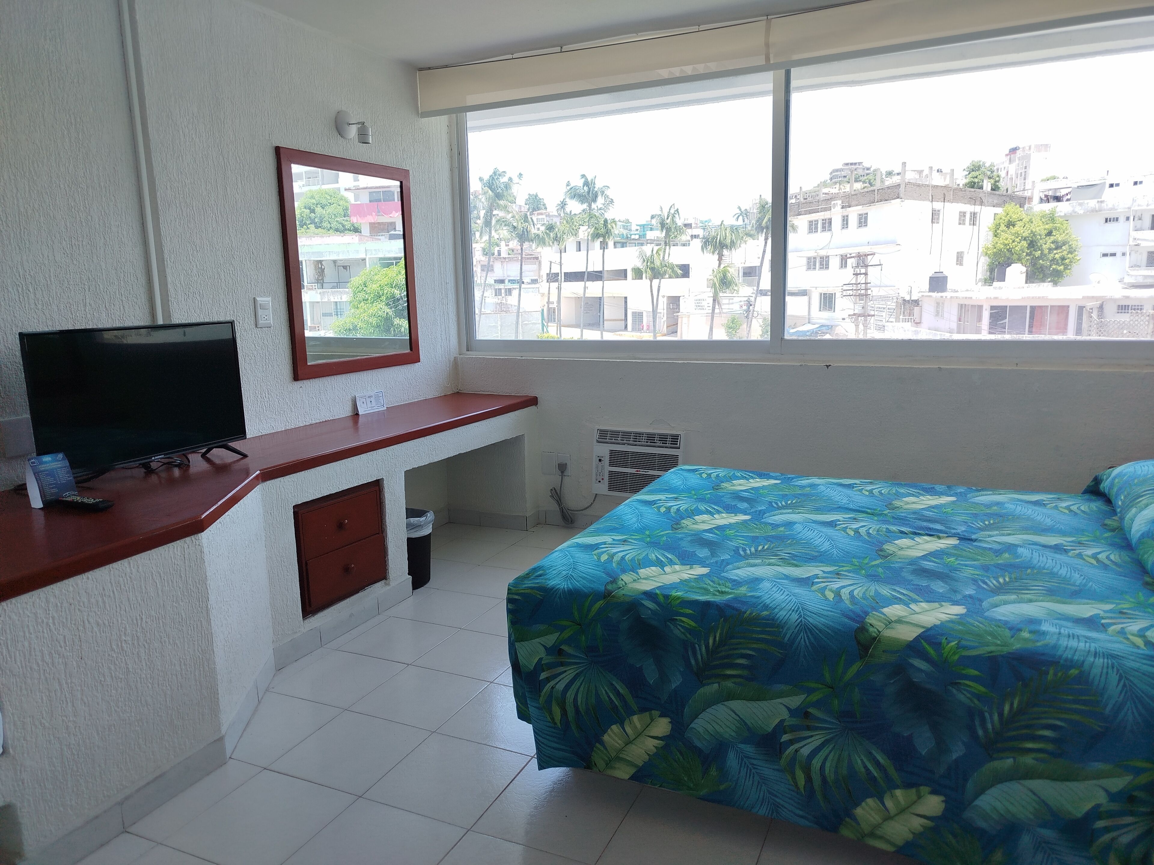 Une chambre d’hôtel avec un lit, un bureau avec une télévision et une fenêtre donnant sur des immeubles et des palmiers.