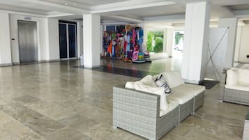 A spacious lobby with a high ceiling, a central seating area with a wicker sofa, a ceiling fan, and a view of a clothing store in the background.