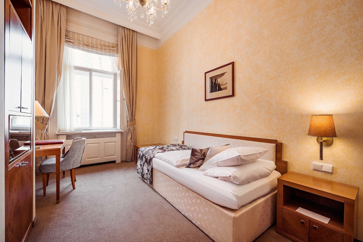Habitación de hotel con cama, escritorio, silla, lámpara y ventana con cortinas.