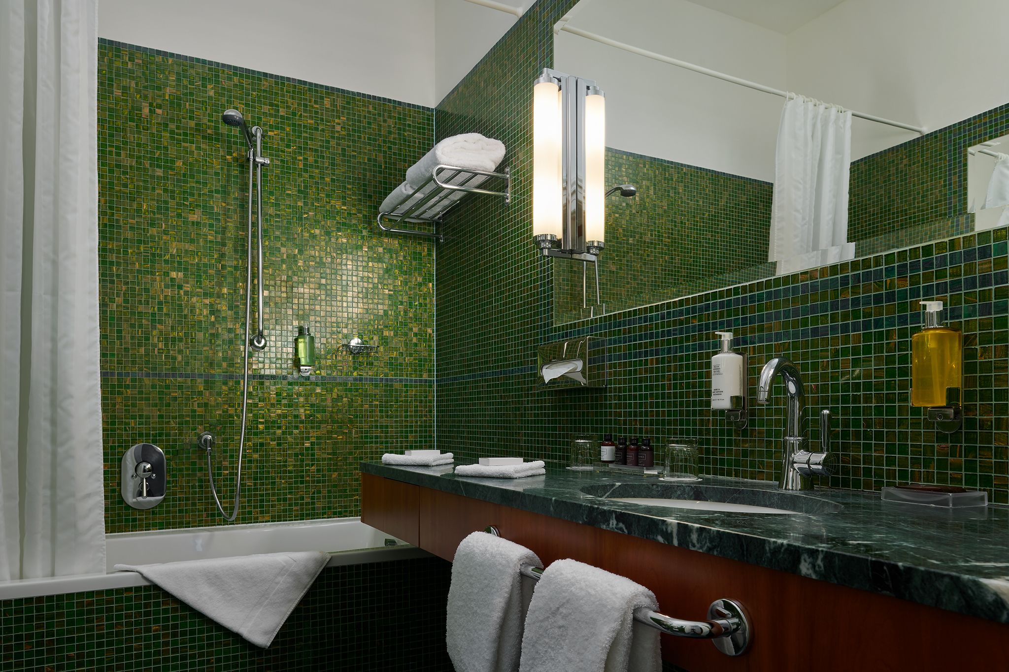 Ein Badezimmer mit grüner Mosaikfliesenwand, einer Dusche, einem Waschbecken mit Spiegel und einem Handtuchhalter.