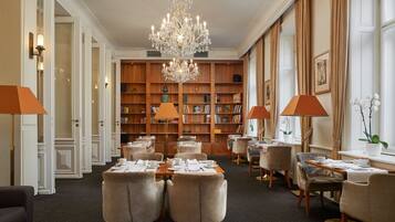 A hotel lobby with a chandelier, bookshelves, and tables set for dining.