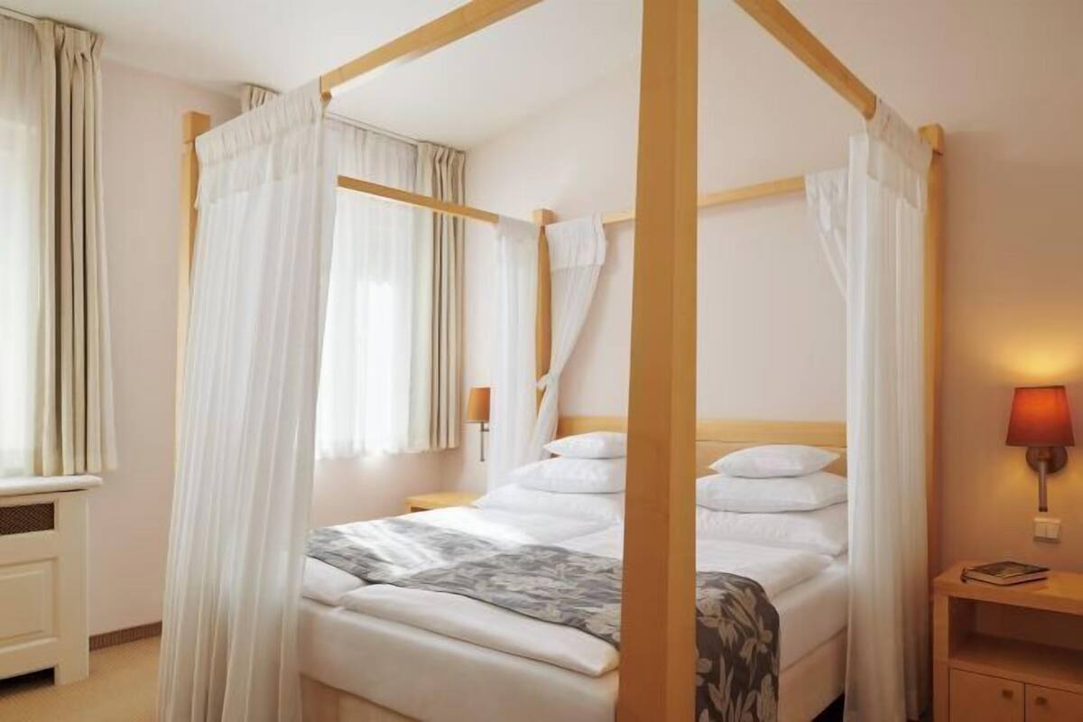 Una cama con dosel con sábanas blancas, una mesita de noche con lámpara y una ventana con cortinas.