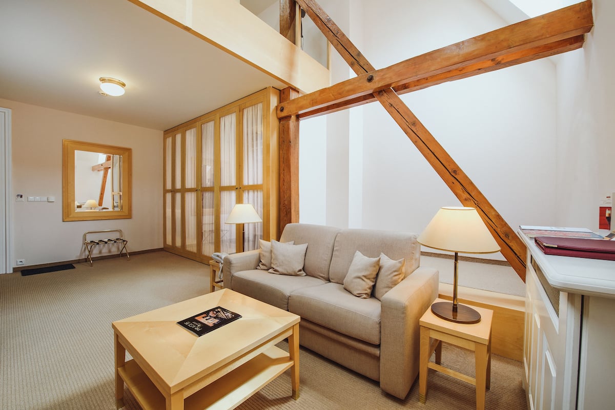 Una sala de estar acogedora con un sofá, una mesa baja, una lámpara y una pequeña mesa con un libro. Se aprecia una estructura de vigas de madera en el techo y una escalera al fondo.