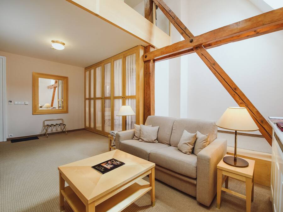 A cozy living room with a sofa, a coffee table, a lamp, and a small table with a book. There is a wooden beam structure on the ceiling and a ladder in the background.