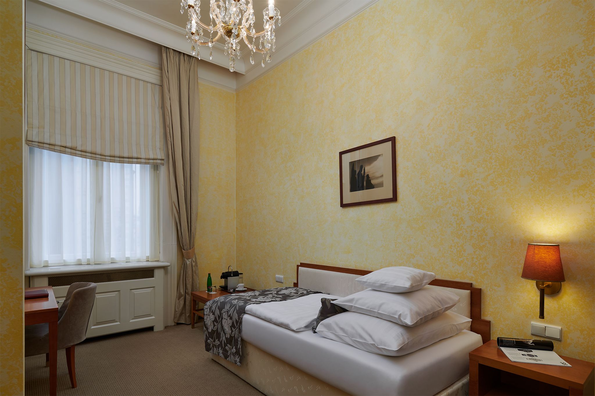 Une chambre d’hôtel avec un lit, une table de chevet, une lampe, une fenêtre avec des rideaux, un radiateur et un tableau au mur.